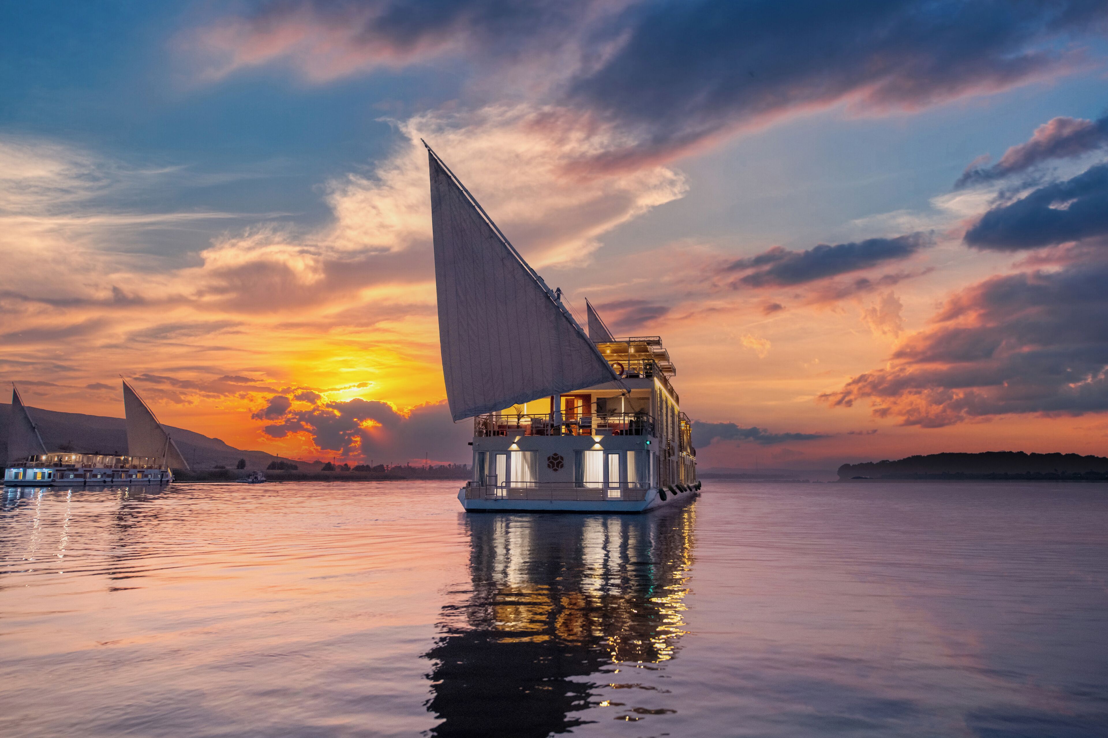 A riverboat with multiple decks and a large sail, surrounded by calm water and a sunset sky.