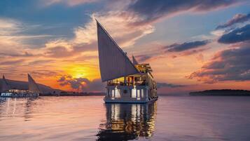 A riverboat with multiple decks and a large sail, surrounded by calm water and a sunset sky.