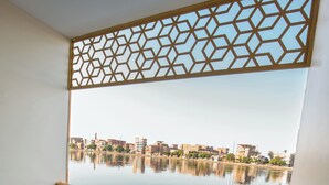 A balcony with a wooden floor, two chairs, a small table, and a view of a calm body of water and buildings across the water.