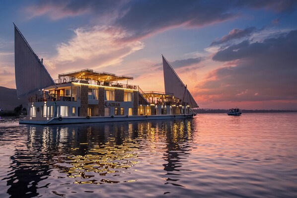 A large, multi-deck houseboat with a prominent sail structure, situated on calm water during sunset. - Azhar Dahabiya Luxury Suites (Esna)
