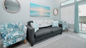 Un salón con un sofá gris, dos sillones estampados, un espejo redondo y una pintura de atardecer en la pared.
