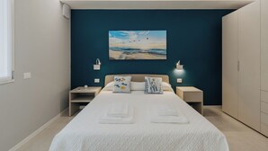 A modern hotel room with a large bed, two bedside tables, a nightstand, a wardrobe, and a painting of a beach scene on the wall. - Modern apartment with pool just steps from the beaches of Lake Garda (Padenghe Sul Garda)