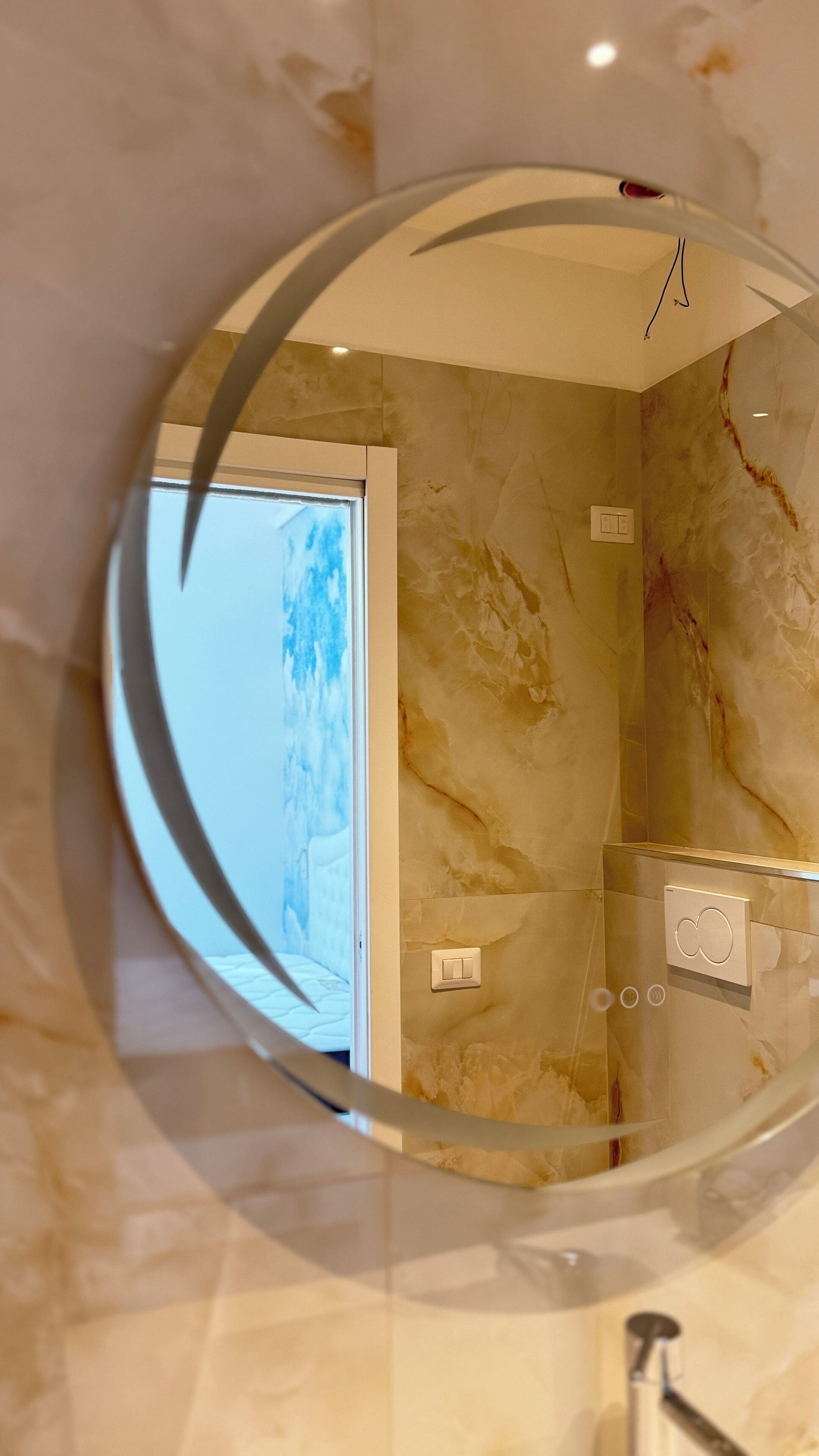 Bagno con pareti in marmo, uno specchio circolare e zona doccia a vista.