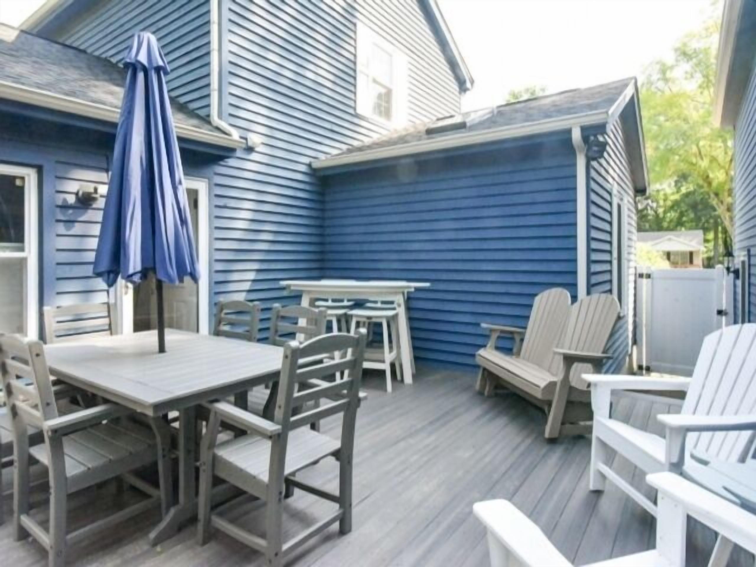 An outdoor patio with a wooden table and chairs, a blue umbrella, and a blue house in the background.