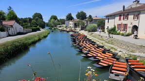 A river with boats lined up on one side, a pedestrian path, and buildings with red shutters. - Holiday Home for 4 "La Marguerite" Countryside Gite near Puy du Fou (Les Herbiers)