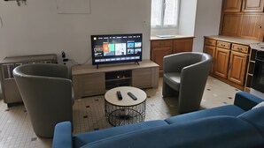 A living room with a TV, two armchairs, a sofa, a small round table, and a kitchen area with wooden cabinets. - Holiday Home for 4 "La Marguerite" Countryside Gite near Puy du Fou (Les Herbiers)