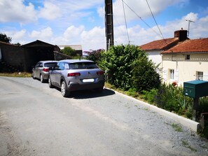 A residential street with two parked cars, a utility box, and a house with a red-tiled roof.