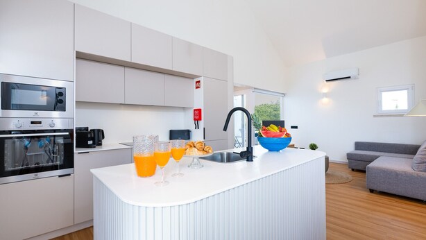 Una cocina moderna con armarios blancos, horno integrado y una isla grande con fregadero. Hay un cuenco de fruta sobre la isla y dos vasos de jugo de naranja en la encimera.