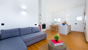 Ein modernes Wohnzimmer mit grauem Sofa, einem kleinen runden Tisch mit Buch und Pflanze, einer Küchenzeile mit Esstisch und Stühlen sowie einer kleinen Kochinsel mit Obstschale.