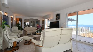 Amplia sala de estar con un sofá grande, un sillón y una mesa de centro. Hay un televisor y un balcón con vistas al mar.