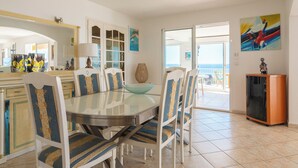 Un comedor con una mesa de vidrio, sillas blancas con tapicería estampada, un cuenco azul y vistas al exterior a través de un ventanal.