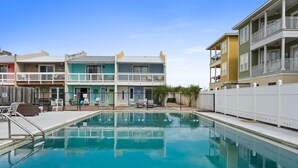 A swimming pool with a white fence, a covered deck, and a row of colorful buildings in the background. - The Pink Flamingo by Book That Condo (Panama City Beach)