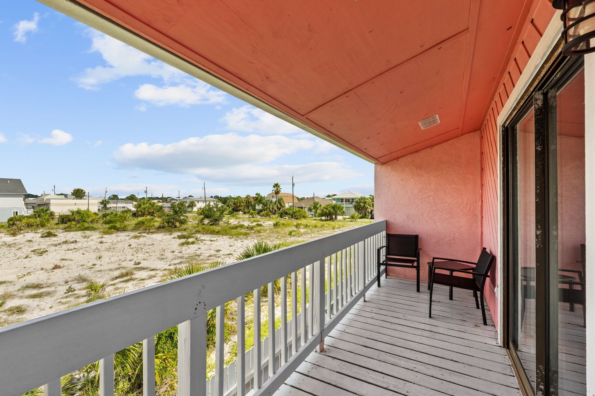 Un balcón con barandilla blanca, piso de madera y vista a una playa arenosa con vegetación escasa y edificios distantes.
