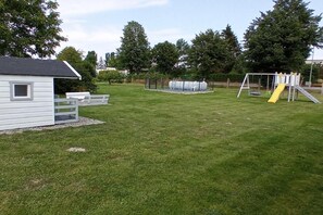 Un espacio exterior con césped, un edificio blanco pequeño, una piscina y juegos infantiles.