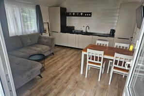 A compact living space with a sofa, a dining table with chairs, a kitchen area with a sink, and a window with curtains. - Family Holiday Homes, Playground, Summer Pool (Ustronie Morskie)