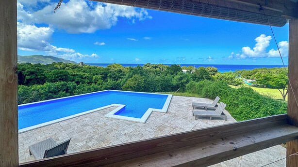 Blick auf einen Swimmingpool mit Holzterrasse und Liegestühlen, mit Blick auf eine üppige grüne Landschaft und den Ozean.