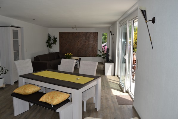 Un salon moderne comprenant une table à manger, un canapé et des fauteuils, une grande fenêtre et une lampe fixée au mur.