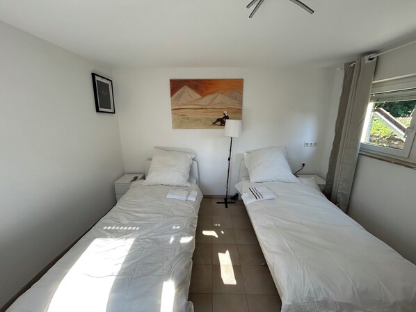 A small, clean room with two single beds, a nightstand, a lamp, and a painting on the wall. - Space for 4 (Unknown)