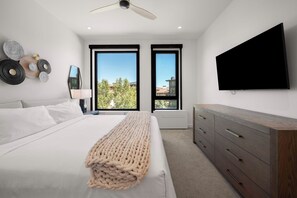 A modern bedroom with a bed, a flat-screen TV, a dresser, and two windows with a view of trees and buildings.