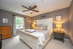 Une chambre à coucher comprenant un lit, des tables de chevet, un ventilateur de plafond, une fenêtre avec des rideaux et des œuvres d’art accrochées aux murs.