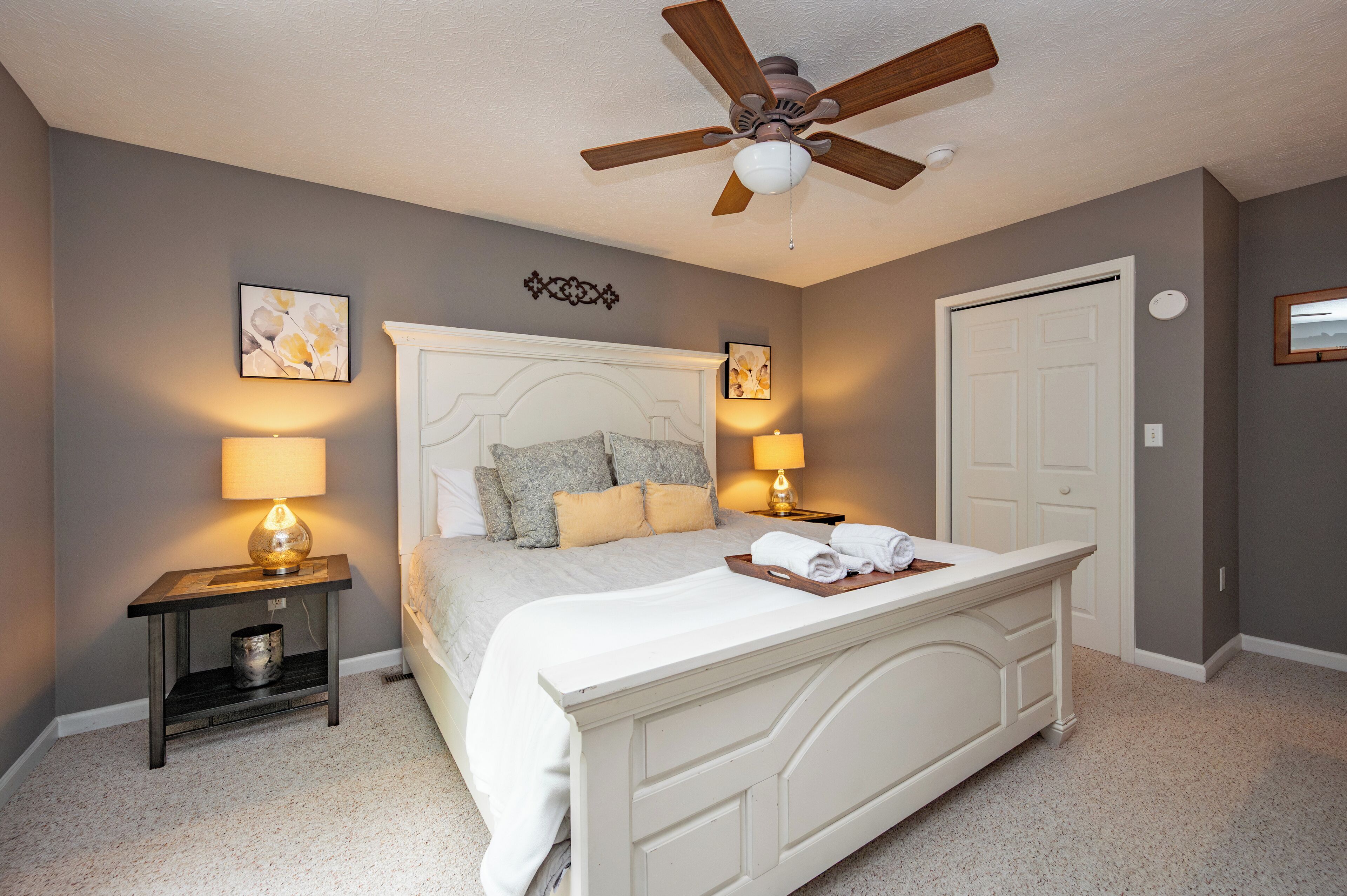 A bedroom with a bed, two bedside tables with lamps, a ceiling fan, and a door.