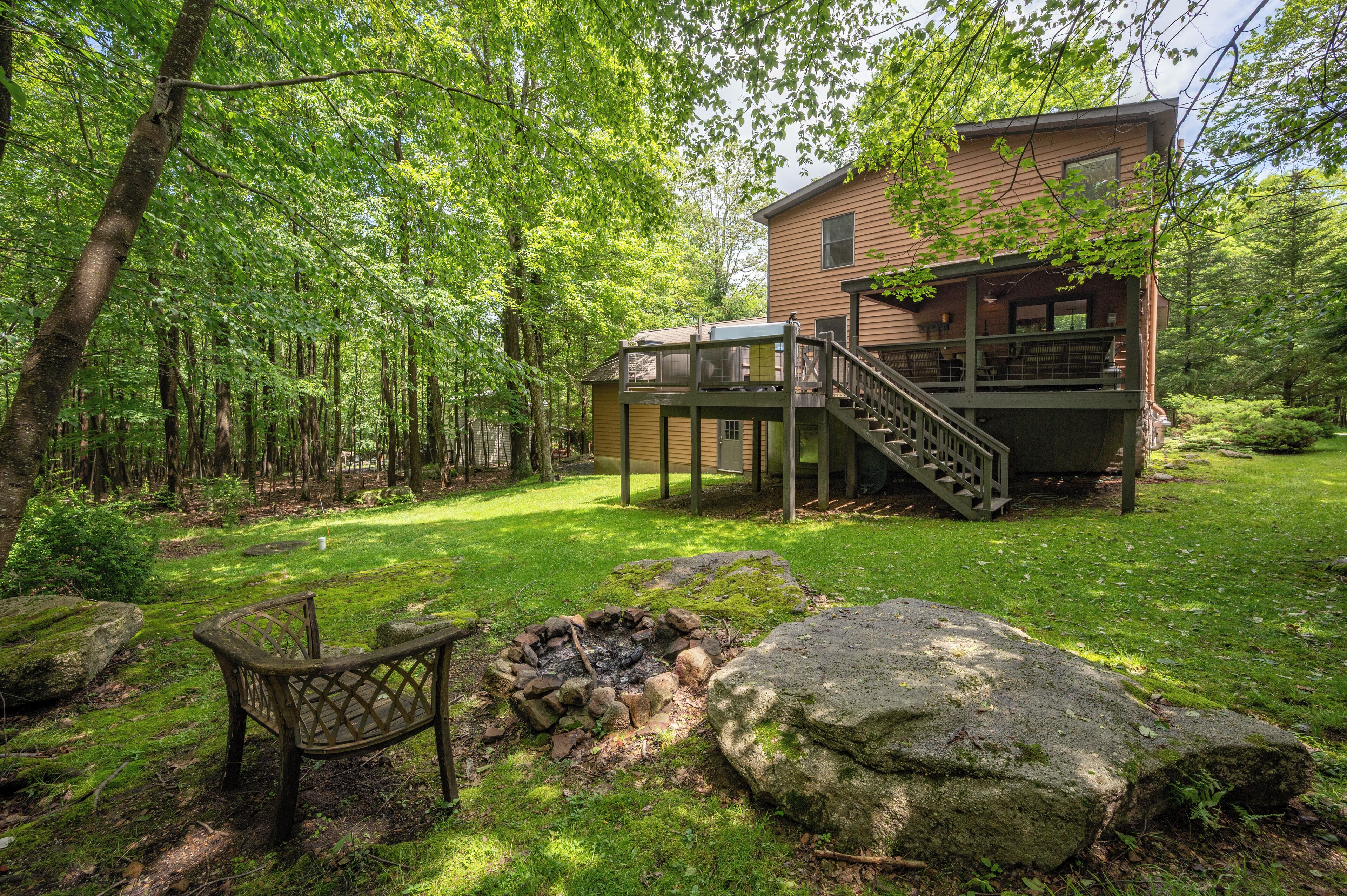 A wooden cabin with a deck and stairs leading to a second floor, surrounded by a forested area with a fire pit and a bench.