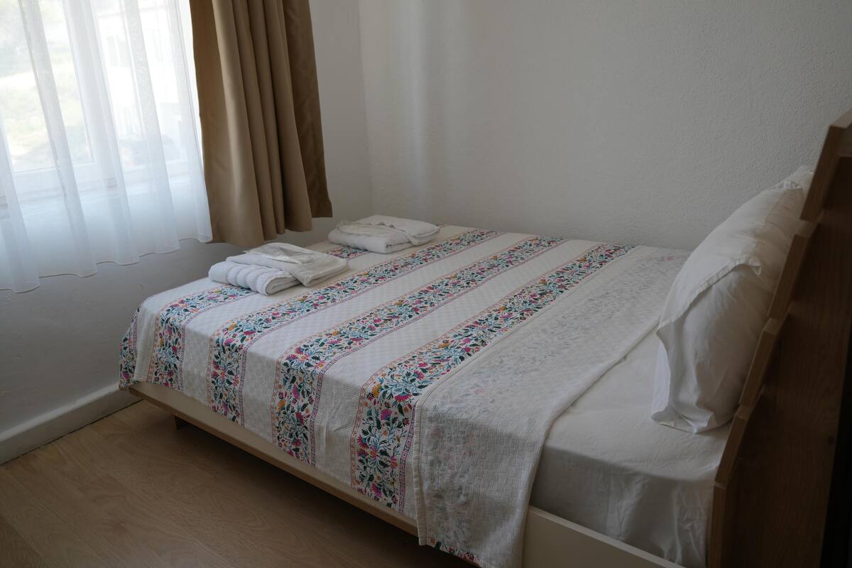 A neatly made bed with a floral bedspread, a pair of folded towels, and a window with sheer curtains.
