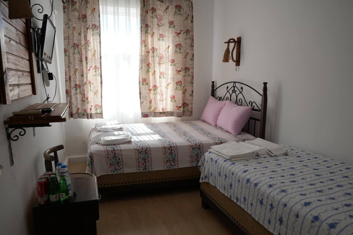 A bedroom with a bed, a desk with a chair, a window with curtains, and a wall-mounted shelf.