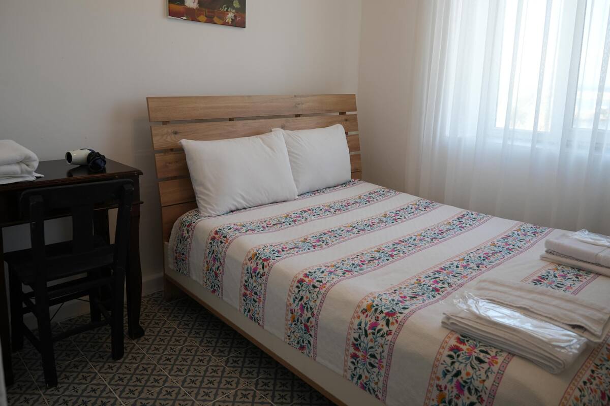 A bedroom with a wooden headboard, a bed with a floral bedspread, a chair, a table with a towel, and a painting on the wall.