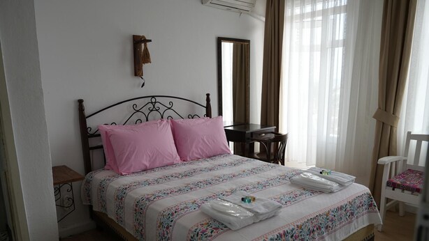 Una cama bien hecha con almohadillas rosas y una colcha estampada, una mesita de noche de madera con una lámpara, un pequeño escritorio con una silla, y una silla blanca con un cojín colorido.