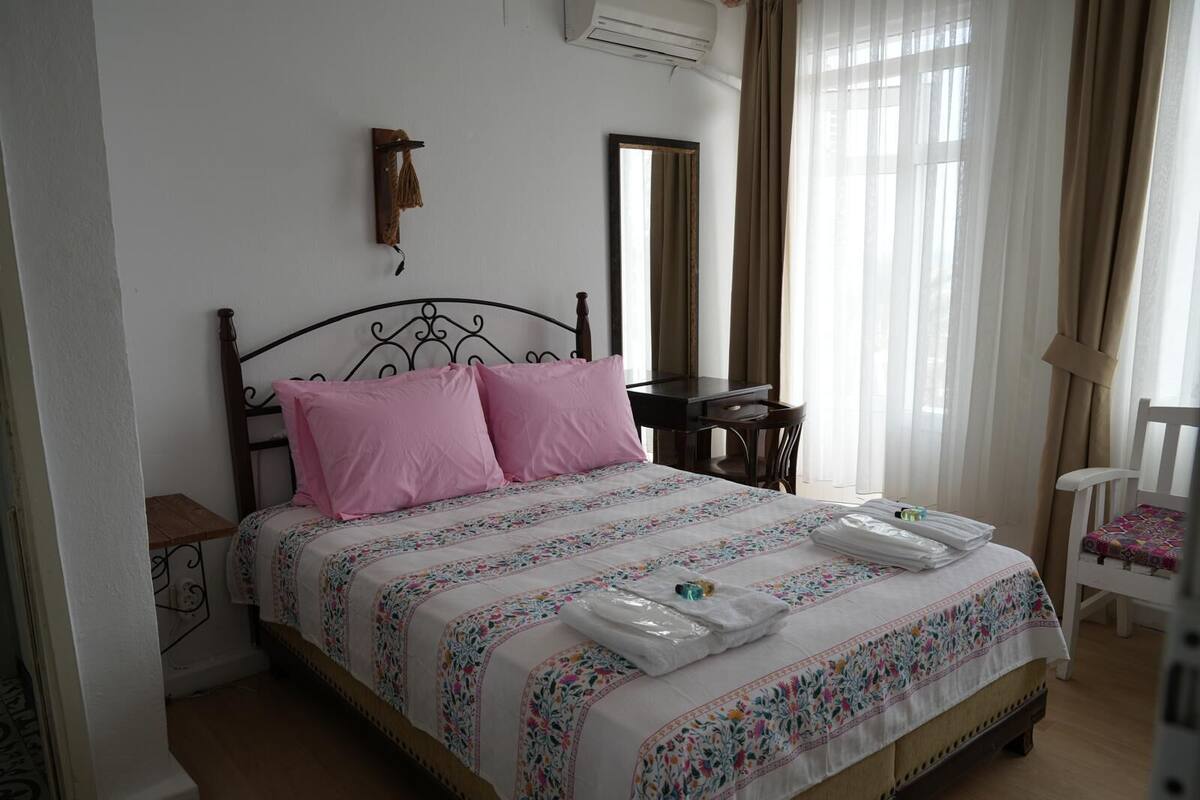 A neatly made bed with pink pillows and a floral bedspread, a wooden nightstand with a lamp, a small desk with a chair, and a white chair with a colorful cushion.