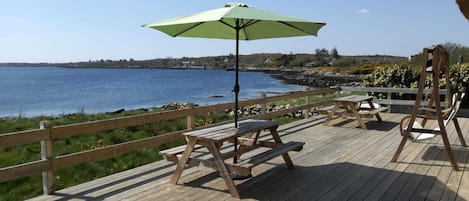 En træterrasse med picnicborde og en parasol med udsigt over vandet.
