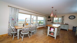 Een eetruimte met een houten tafel, witte stoelen en uitzicht op zee door grote ramen.