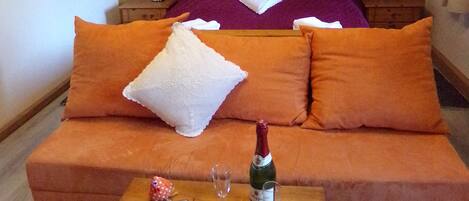 Habitación de hotel con cama, almohadas anaranjadas y blancas, una mesa de madera con una botella de champán, dos copas y un teléfono inteligente.