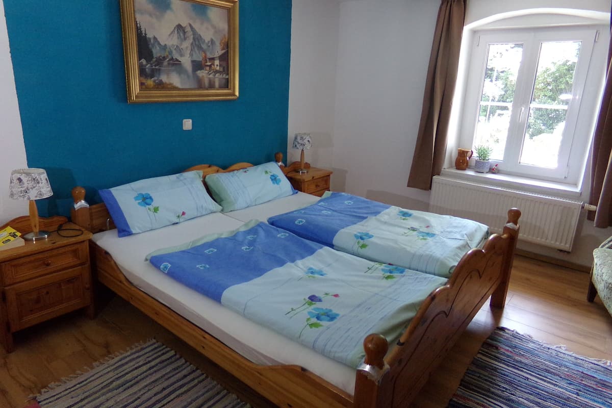 Une chambre à coucher avec un lit, une tête de lit en bois, des tables de chevet, une lampe, un tableau, une fenêtre avec des rideaux et un radiateur.