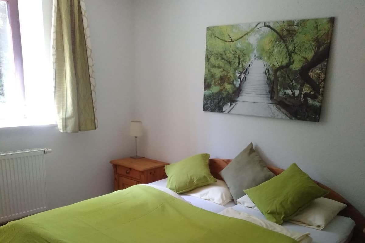 Une chambre à coucher avec un lit, une table de chevet, une lampe, une fenêtre avec des rideaux et un tableau au mur.
