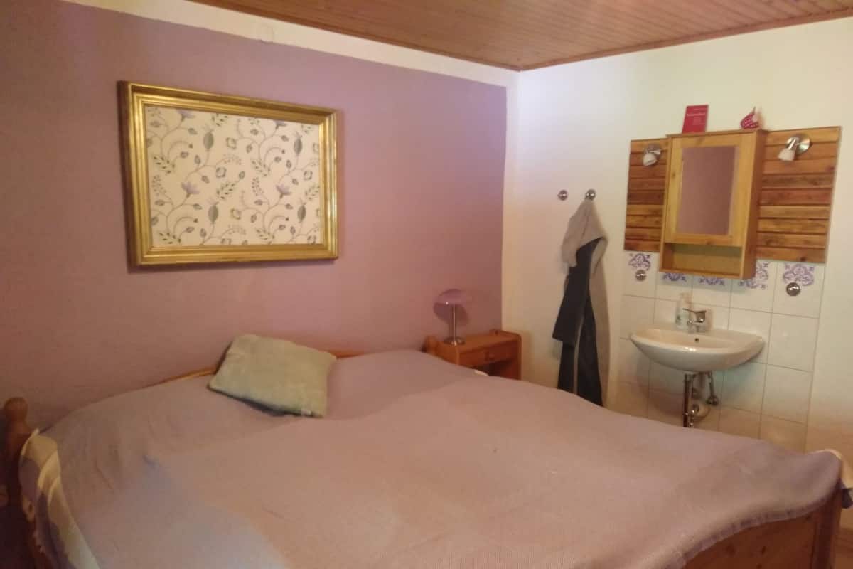 Une chambre à coucher comprenant un lit, une table de chevet, une lampe de chevet, un tableau floral encadré et une commode en bois dotée d’un miroir et d’un lavabo.
