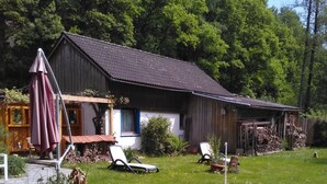 Een rustiek houten huis met pannendak, omgeven door een weelderige tuin met gele bloemen en een verhard terras met vuurkorf.