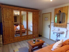 Um quarto com guarda-roupa de madeira, uma cama, uma mesa de madeira com uma garrafa e copos, uma pia e um espelho.