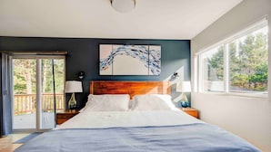 Une chambre à coucher avec un grand lit, une tête de lit en bois, un tableau accroché au mur et une vue sur les arbres par la fenêtre.
