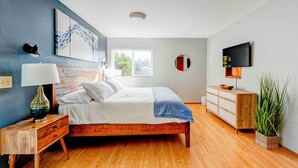 Une chambre à coucher moderne avec un lit en bois, une table de chevet avec une lampe, une commode, un téléviseur fixé au mur et une fenêtre donnant sur des arbres.
