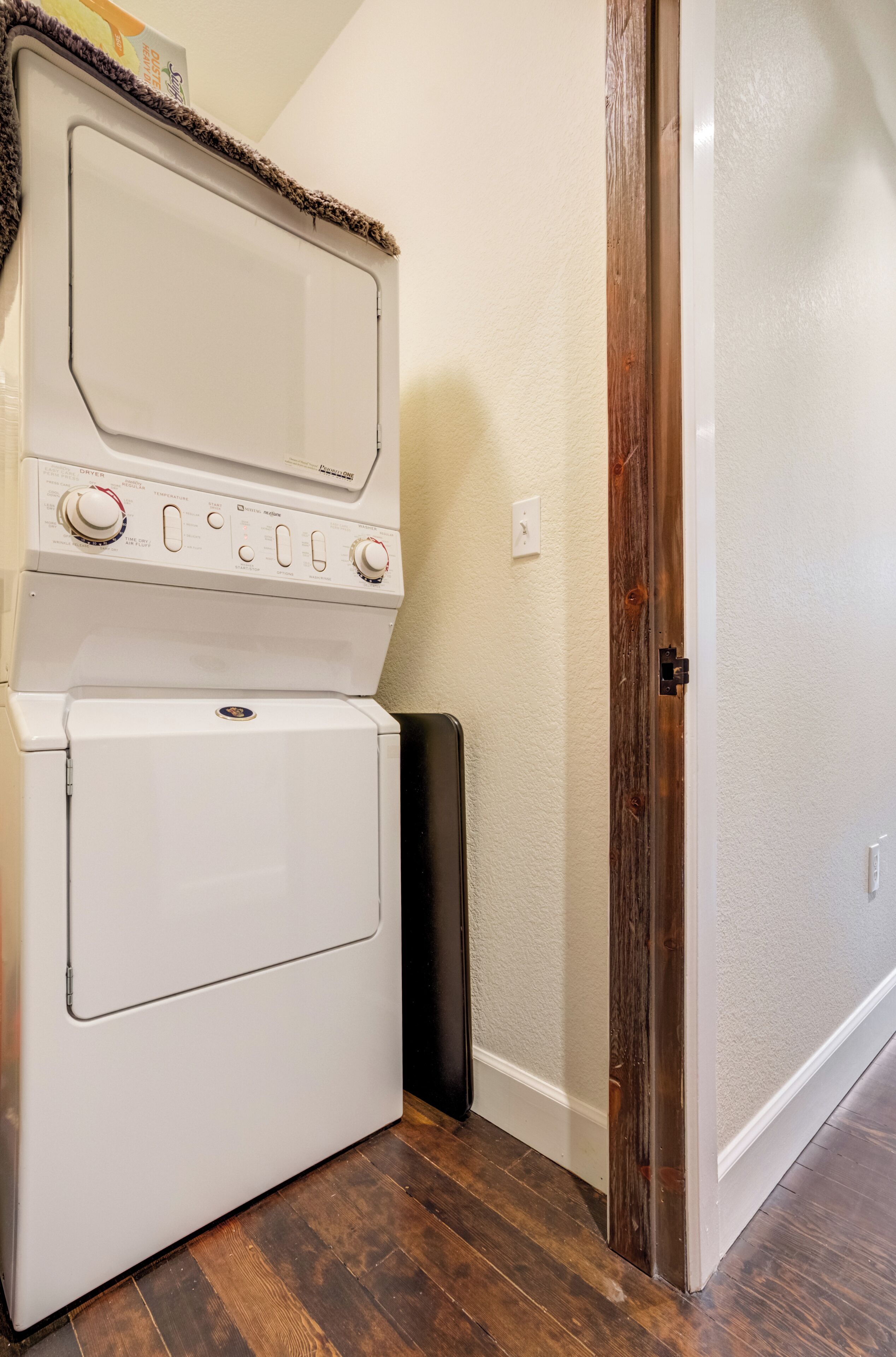 A front-loading washer and dryer in a corner with a wooden door frame and a white wall.