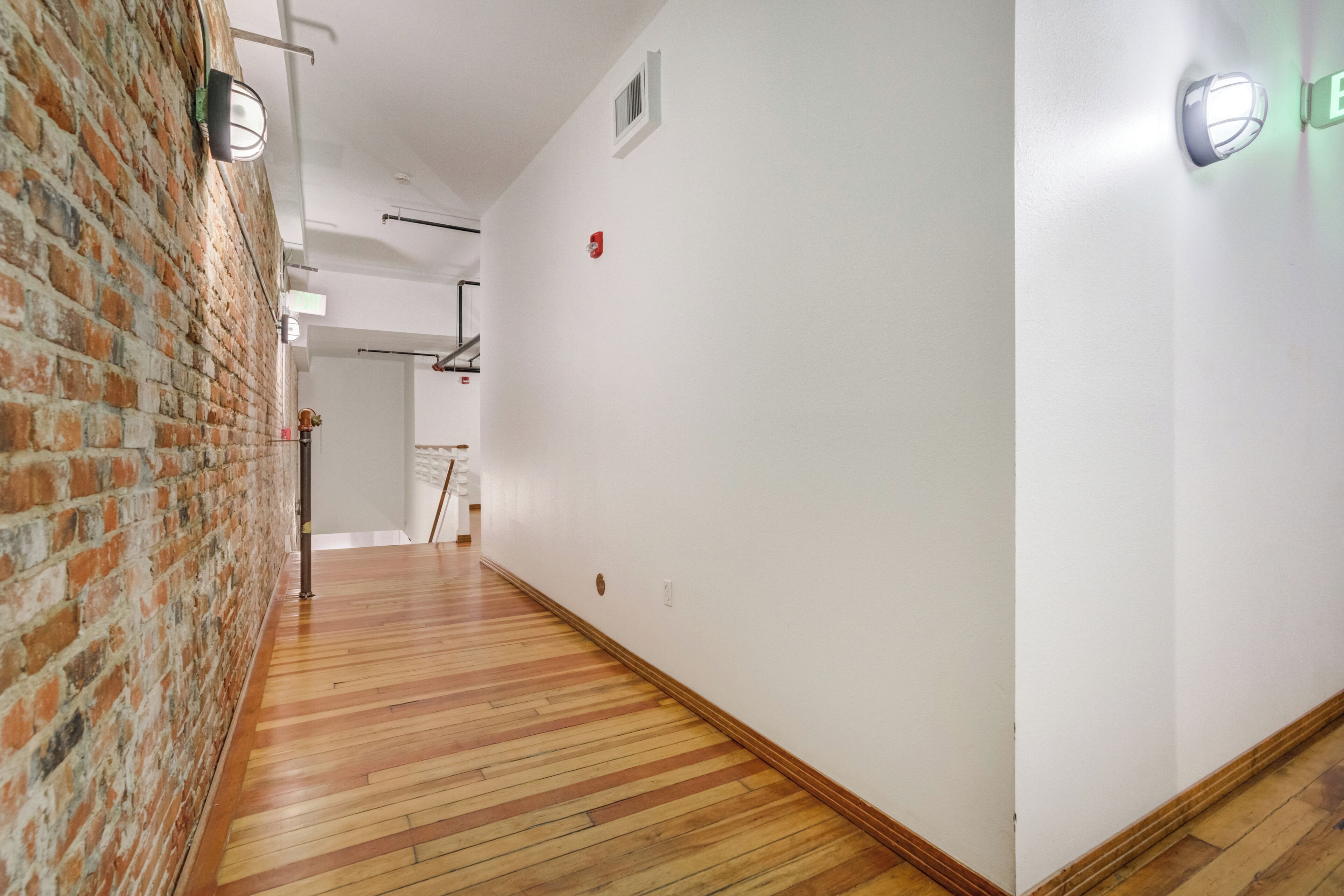 A hallway with a brick wall on one side and a white wall on the other, wooden flooring, and wall-mounted lighting fixtures.