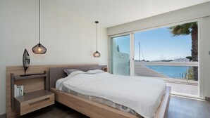 A modern bedroom with a wooden bed, a nightstand, and a large window offering a view of the sea. - Villa in Le Grau du Roi with Stunning Sea Views (Le Grau-du-Roi)