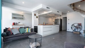A modern apartment with a grey sofa, a small round table, a kitchen with white cabinets, and a staircase. - Villa in Le Grau du Roi with Stunning Sea Views (Le Grau-du-Roi)