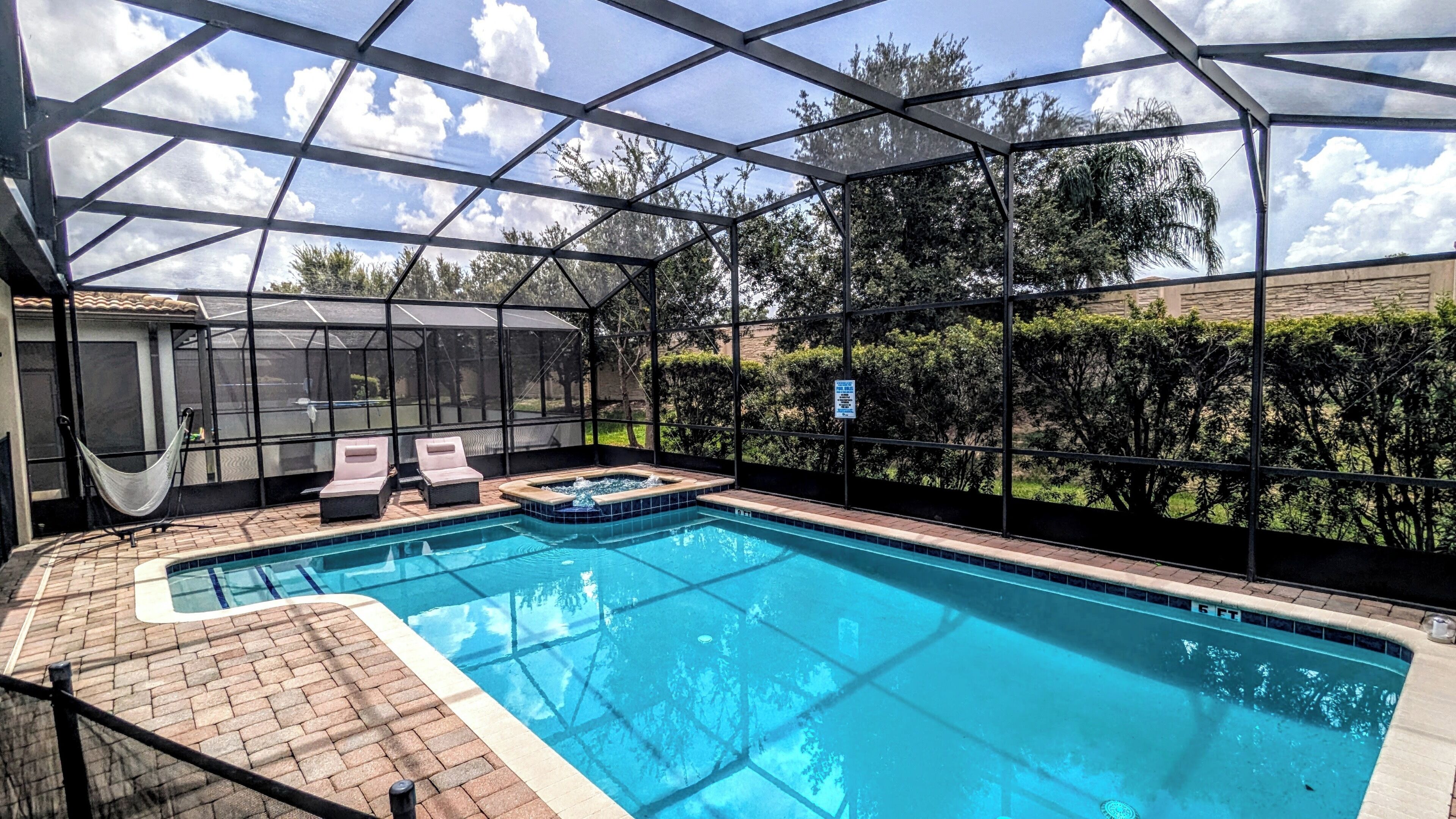 A covered swimming pool with two lounge chairs and a hammock, surrounded by a metal frame and glass panels.