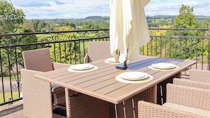 Un balcon agrémenté de meubles en osier, une table dressée avec de l’argenterie et un parasol blanc, et une vue panoramique sur des collines verdoyantes et des arbres.