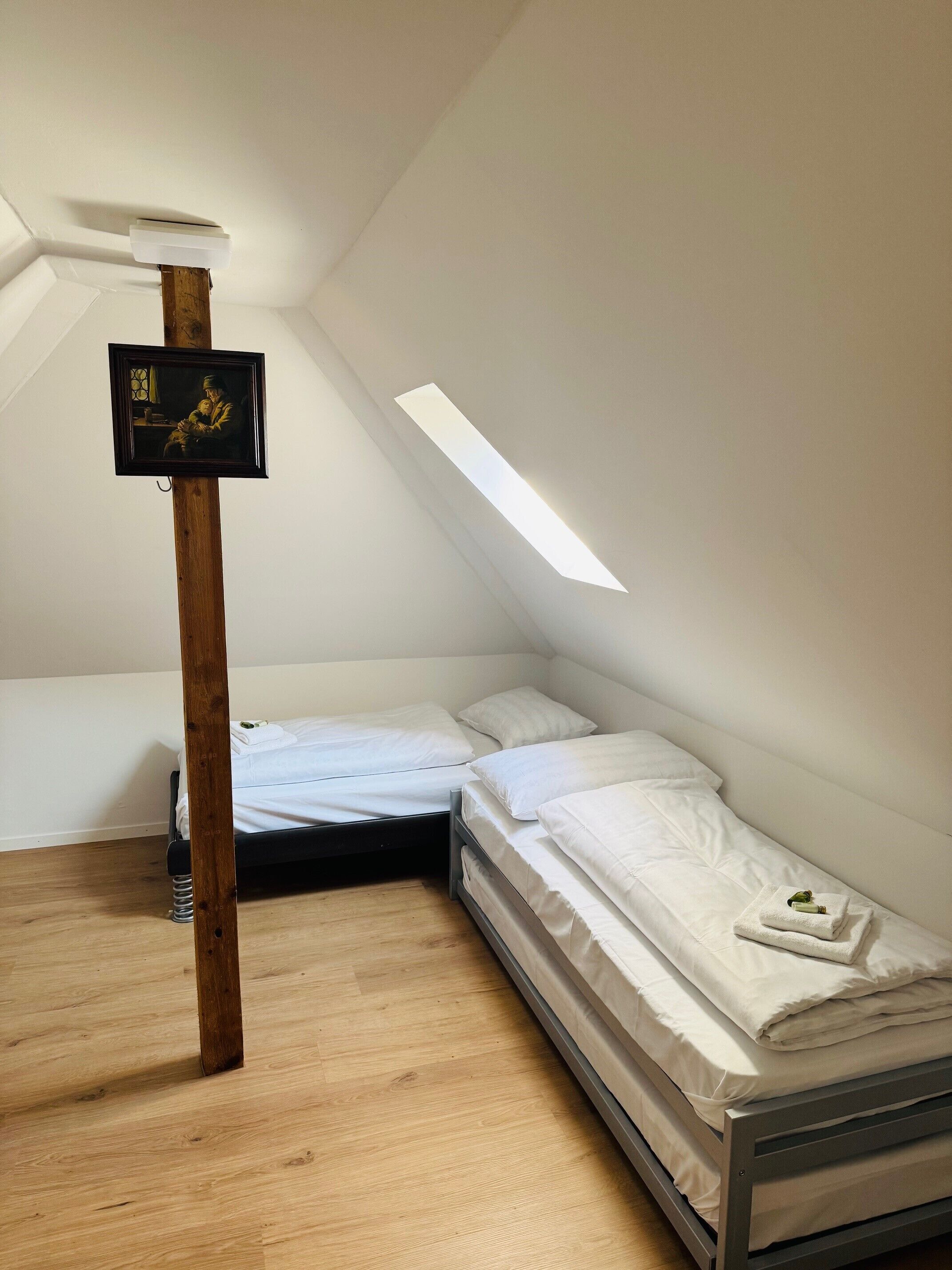 A small, well-lit attic bedroom with a sloped ceiling, two beds, and a wooden beam structure.