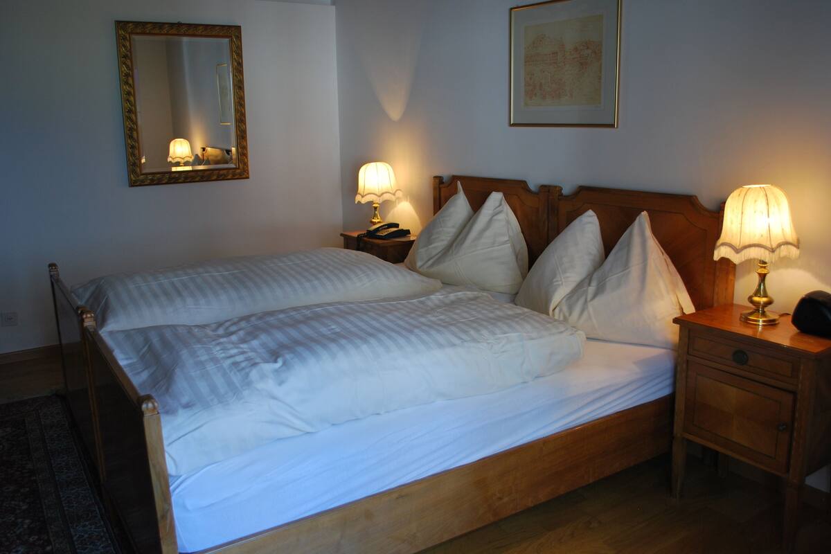 A neatly made bed with white linens and pillows, a wooden headboard, two bedside lamps, a nightstand with a telephone, and a framed picture on the wall.