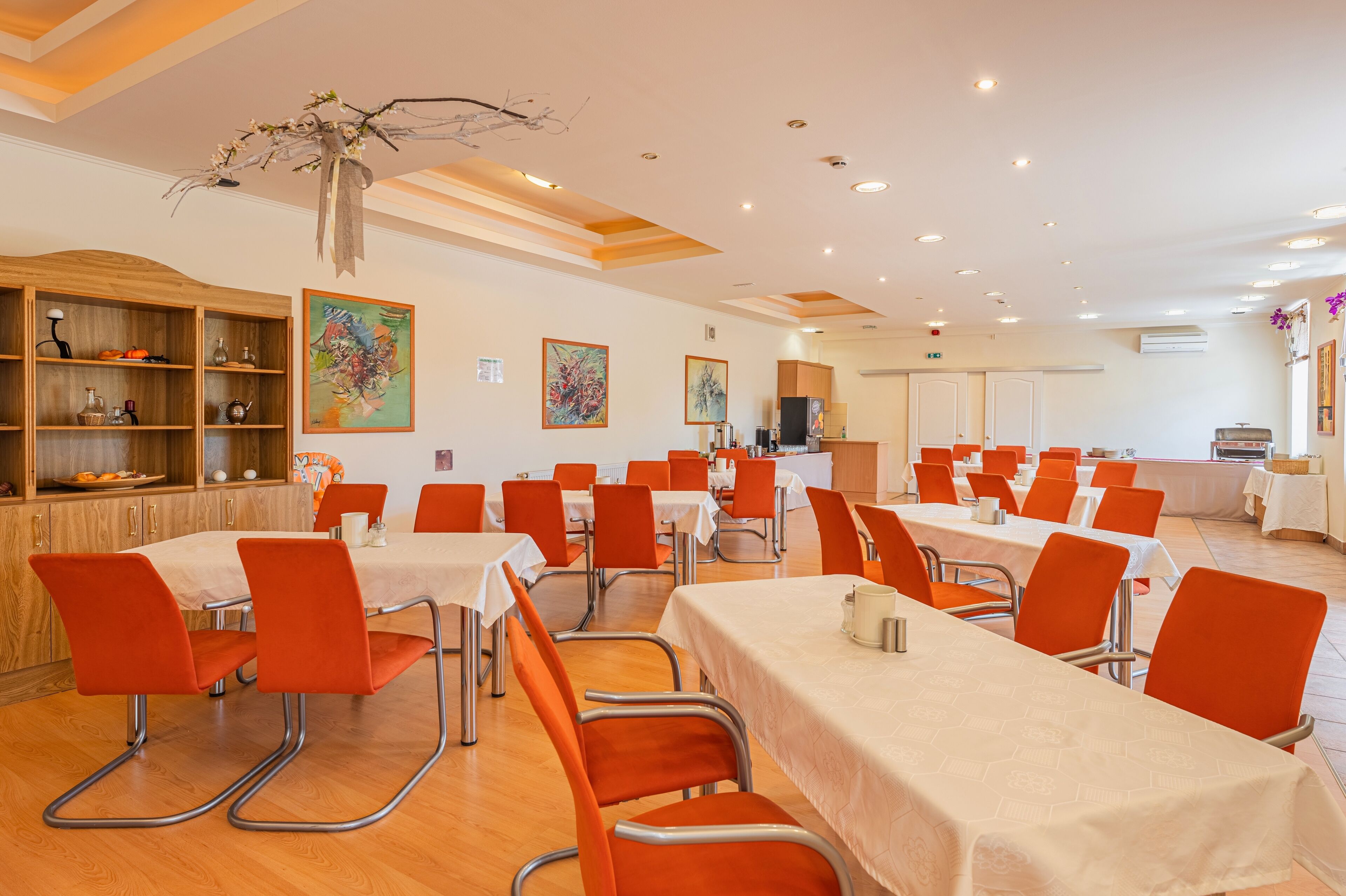 A dining area with tables and chairs, a wooden cabinet with dishes, and artwork on the walls.